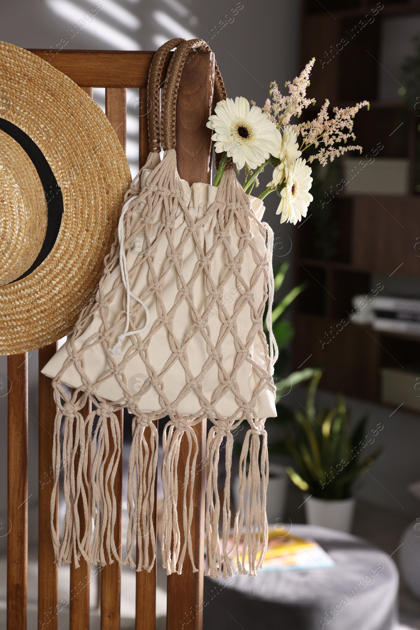 Photo of Handmade macrame shopping bag with flowers and straw hat on wooden chair indoors