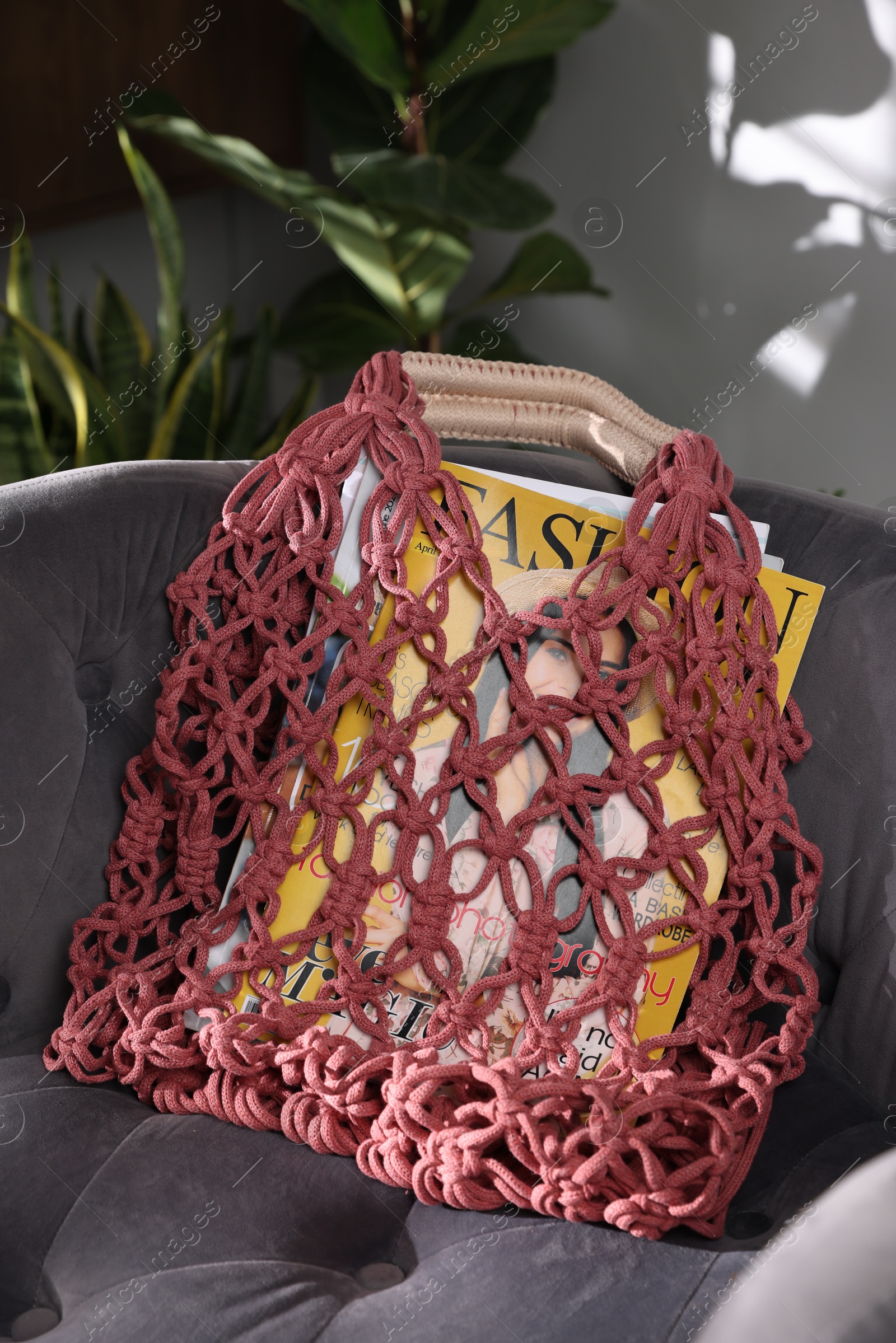 Photo of Handmade macrame shopping bag with magazines on armchair indoors