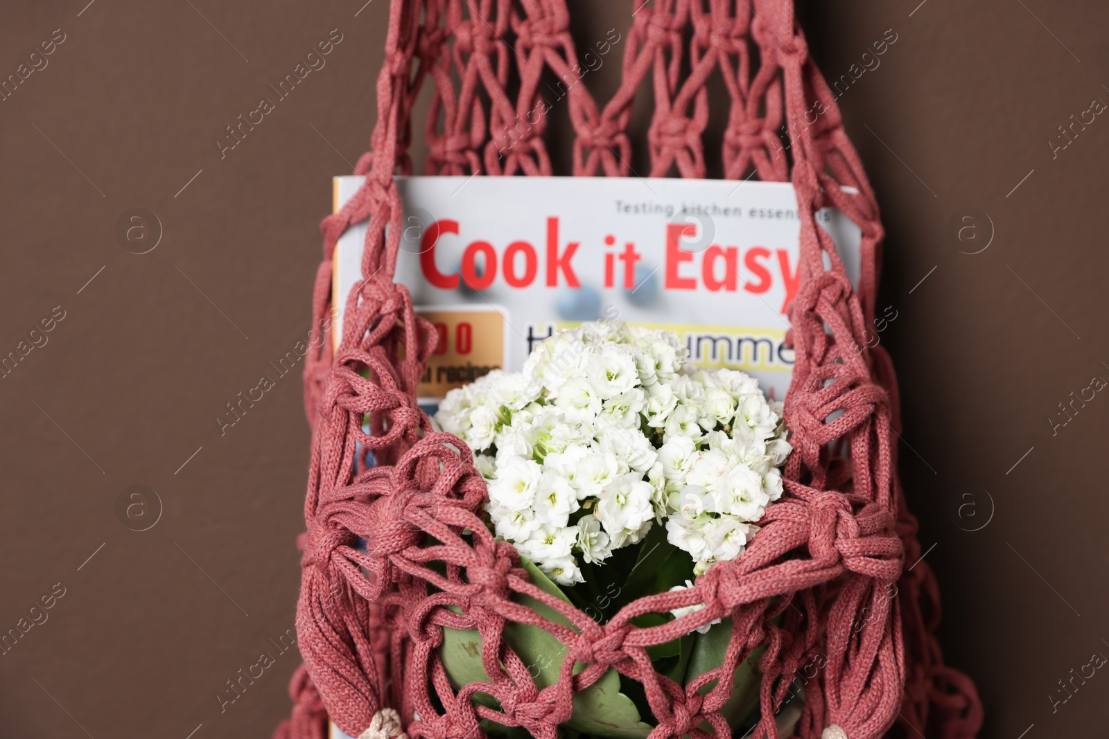 Photo of Handmade macrame shopping bag with magazine and flowers on brown wall, closeup