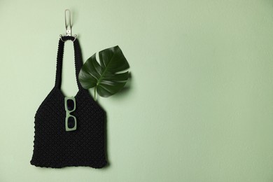 Photo of Handmade macrame shopping bag with leaf and sunglasses on light green wall, space for text