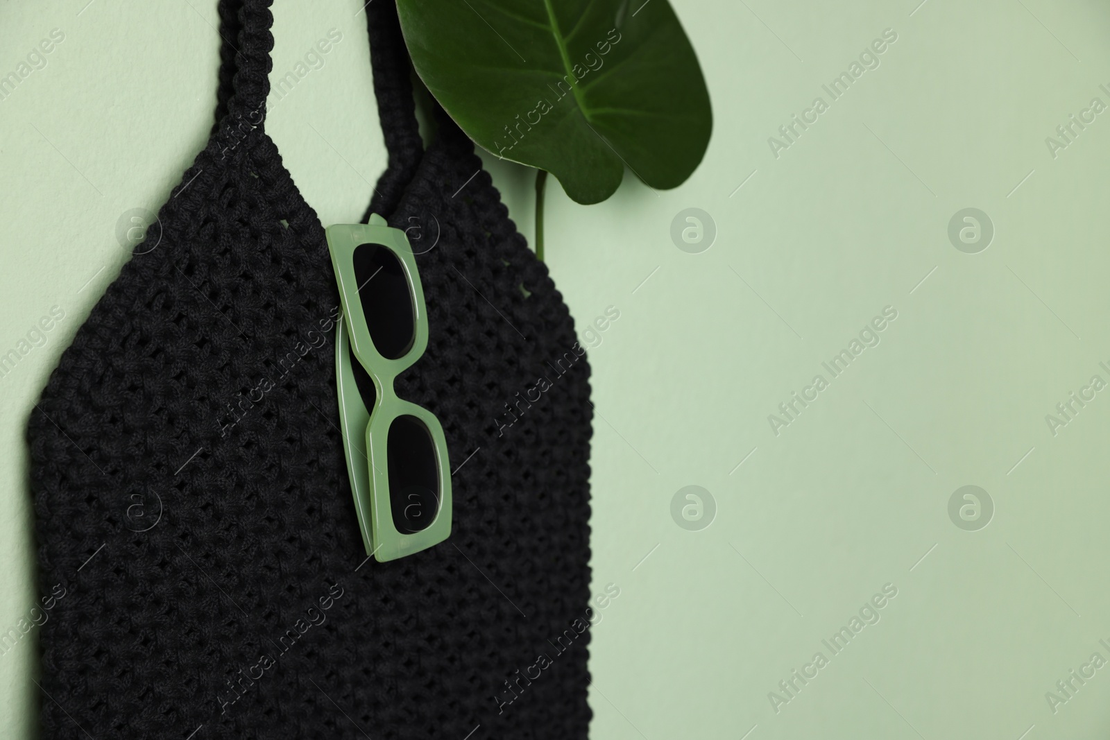 Photo of Handmade macrame shopping bag with leaf and sunglasses on light green wall. Space for text