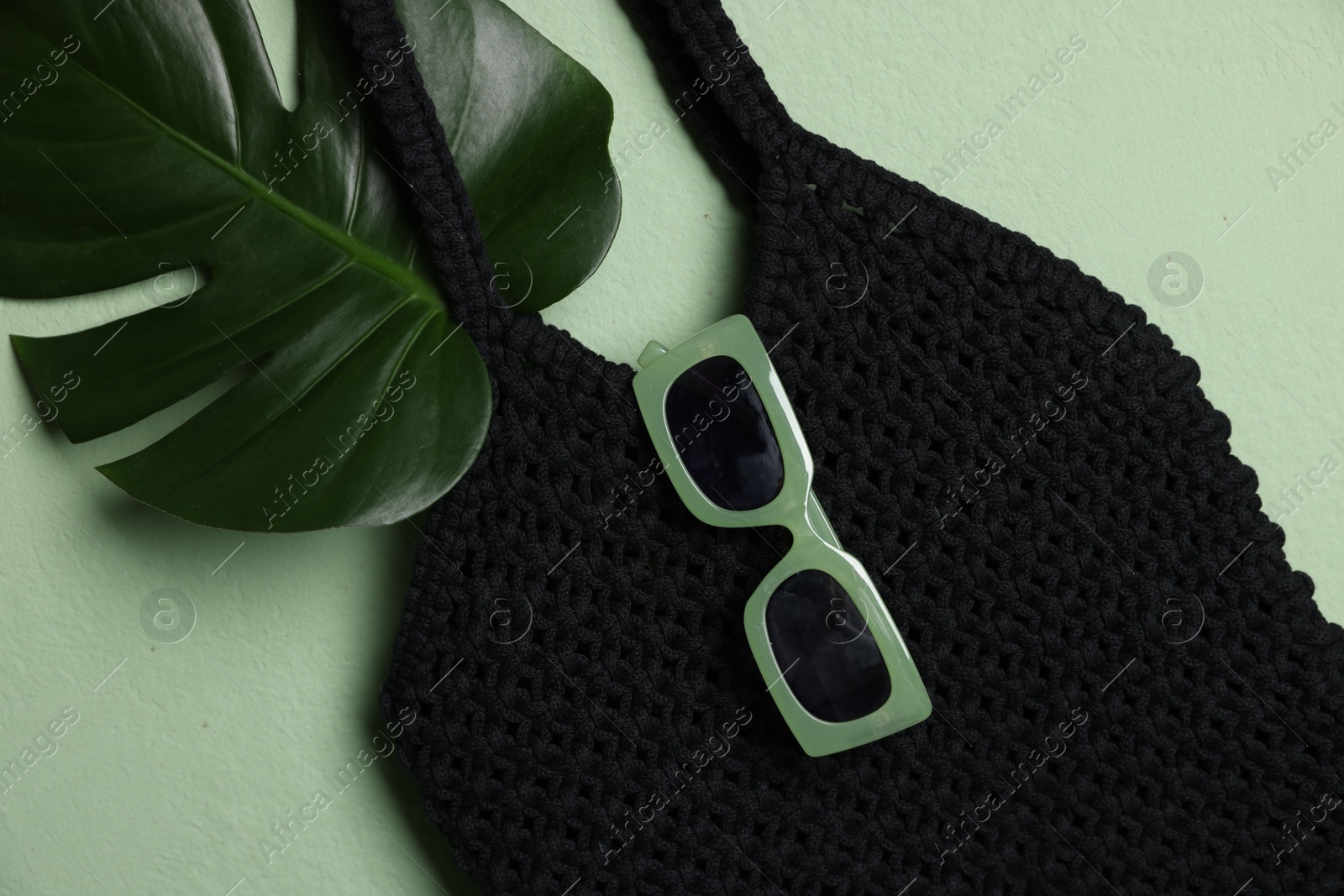 Photo of Handmade macrame shopping bag with leaf and sunglasses on light green background, top view