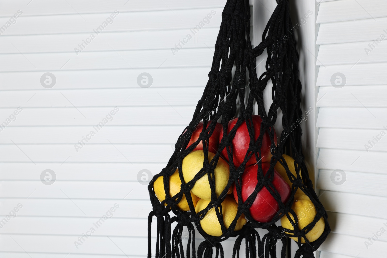 Photo of Handmade macrame shopping bag with fruits on white wall, space for text