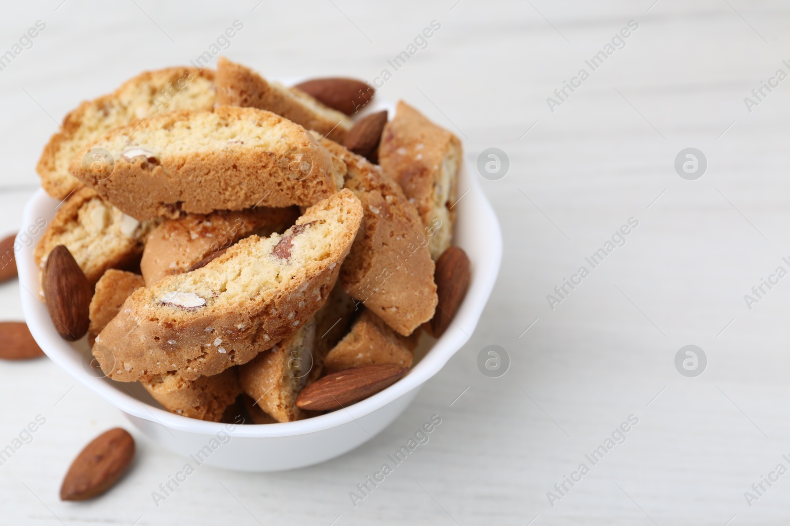 Photo of Tasty almond biscuits (Cantuccini) and nuts in bowl on white wooden table, closeup. Space for text