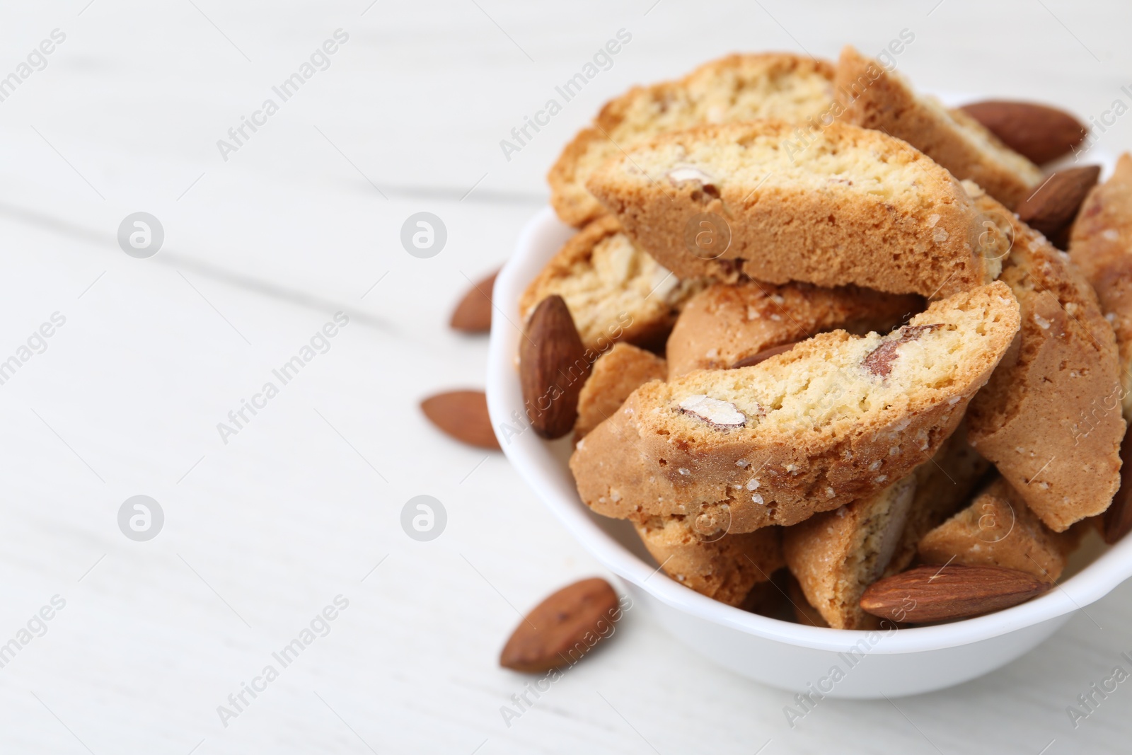 Photo of Tasty almond biscuits (Cantuccini) and nuts in bowl on white wooden table, closeup. Space for text