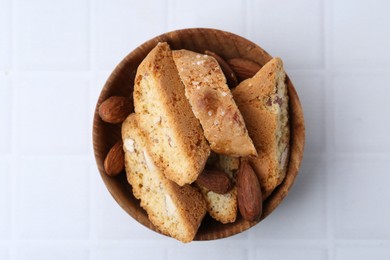 Photo of Tasty almond biscuits (Cantuccini) and nuts in bowl on white tiled table, top view