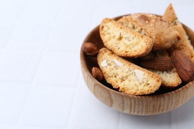 Photo of Tasty almond biscuits (Cantuccini) and nuts in bowl on white tiled table, closeup. Space for text