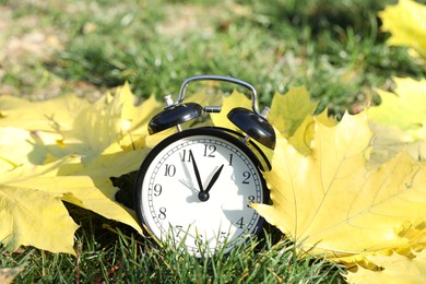 Photo of Autumn time. Alarm clock among fallen leaves on green grass, closeup