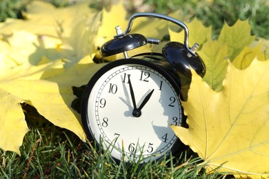 Photo of Autumn time. Alarm clock among fallen leaves on green grass, closeup