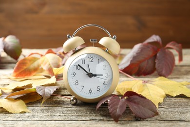 Photo of Autumn time. Alarm clock among leaves on wooden table, closeup