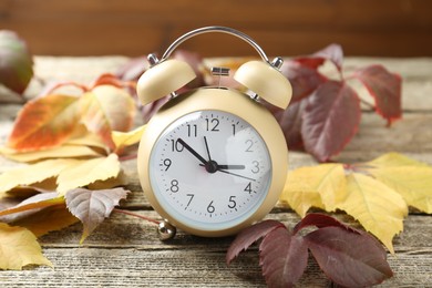 Photo of Autumn time. Alarm clock among leaves on wooden table, closeup