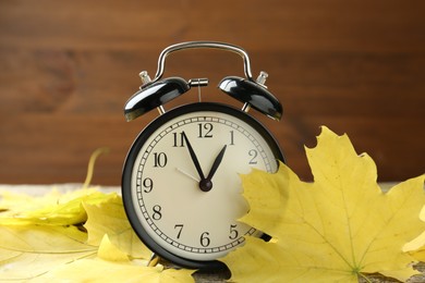 Photo of Autumn time. Alarm clock and leaves on wooden table, closeup