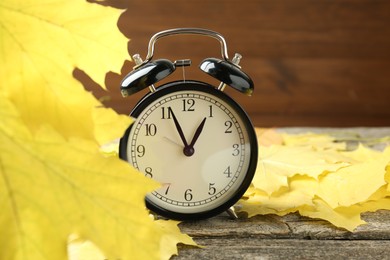 Photo of Autumn time. Alarm clock and leaves on wooden table, closeup