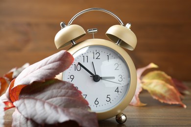 Photo of Autumn time. Alarm clock among leaves on wooden table, closeup