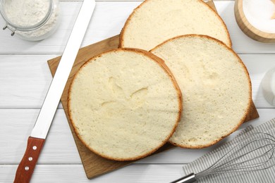 Photo of Delicious cut sponge cake, ingredients and knife on white wooden table, flat lay