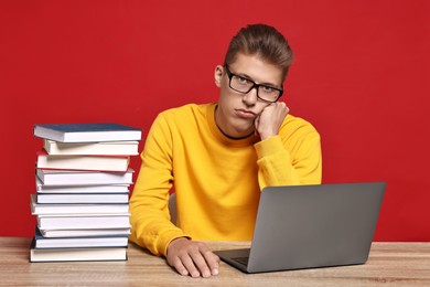 Photo of Tired student before exam with stack of books and laptop at wooden table against red background