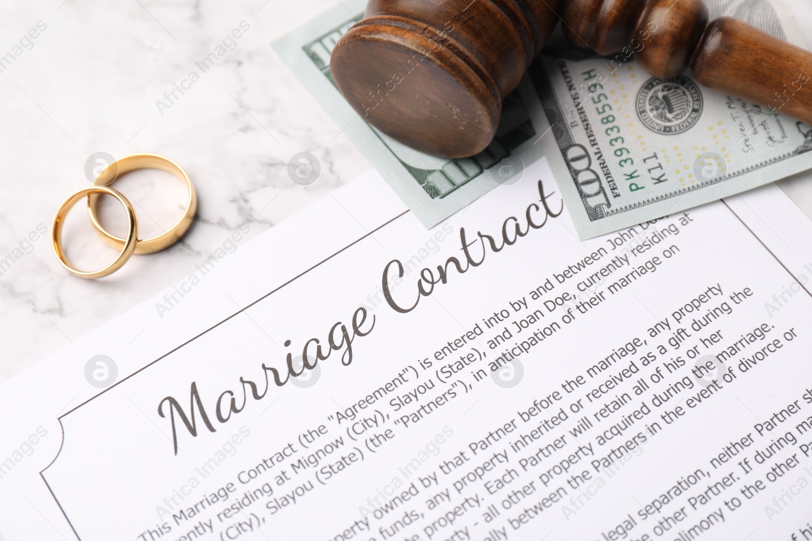 Photo of Marriage contract, dollar bill, gavel and golden rings on white marble table, closeup