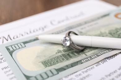 Photo of Marriage contract, dollar bill, pen and ring on wooden table, closeup