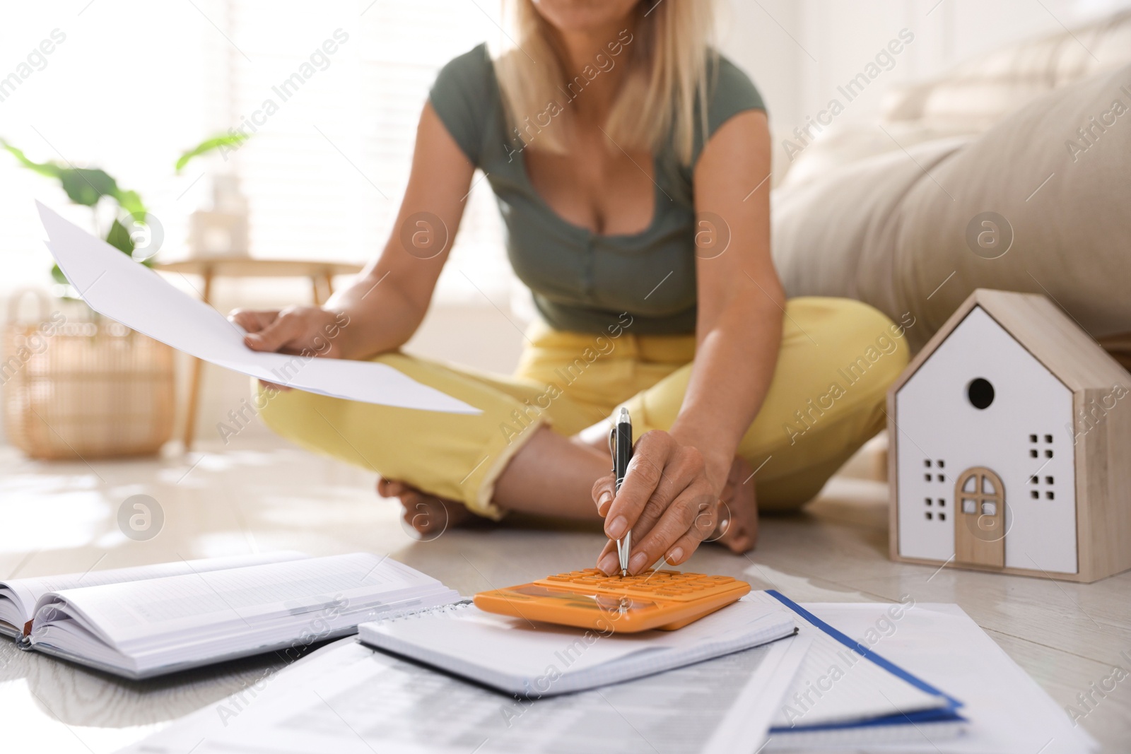 Photo of Budget planning. Woman with notebook using calculator on floor indoors, closeup