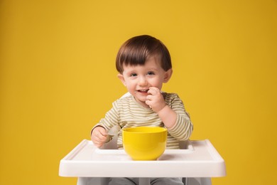 Photo of Cute little kid eating healthy baby food from bowl in high chair on yellow background