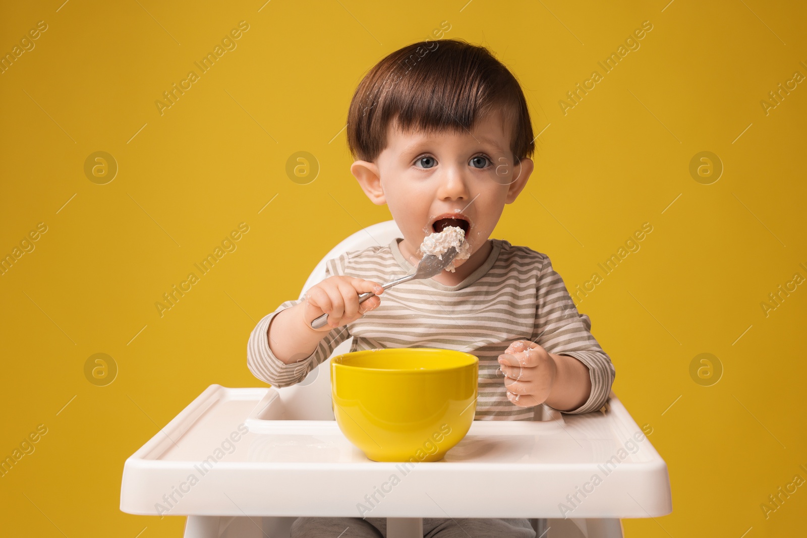 Photo of Cute little kid eating healthy baby food from bowl in high chair on yellow background