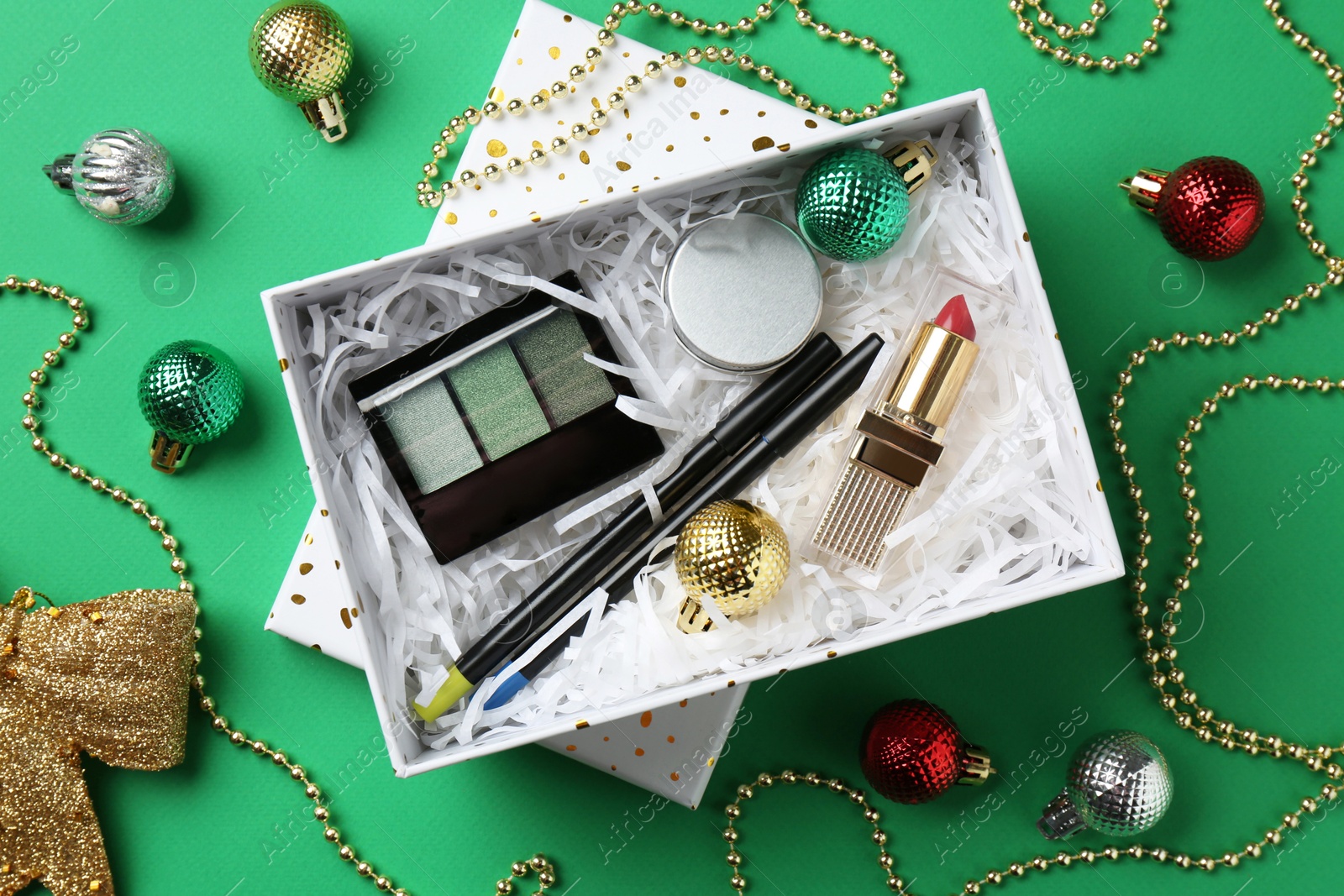 Photo of Christmas gift box with decorative cosmetics and festive ornaments on green background, flat lay