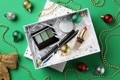 Christmas gift box with decorative cosmetics and festive ornaments on green background, flat lay