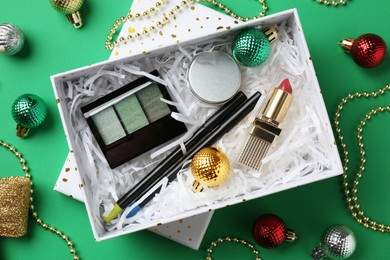 Christmas gift box with decorative cosmetics and festive ornaments on green background, flat lay