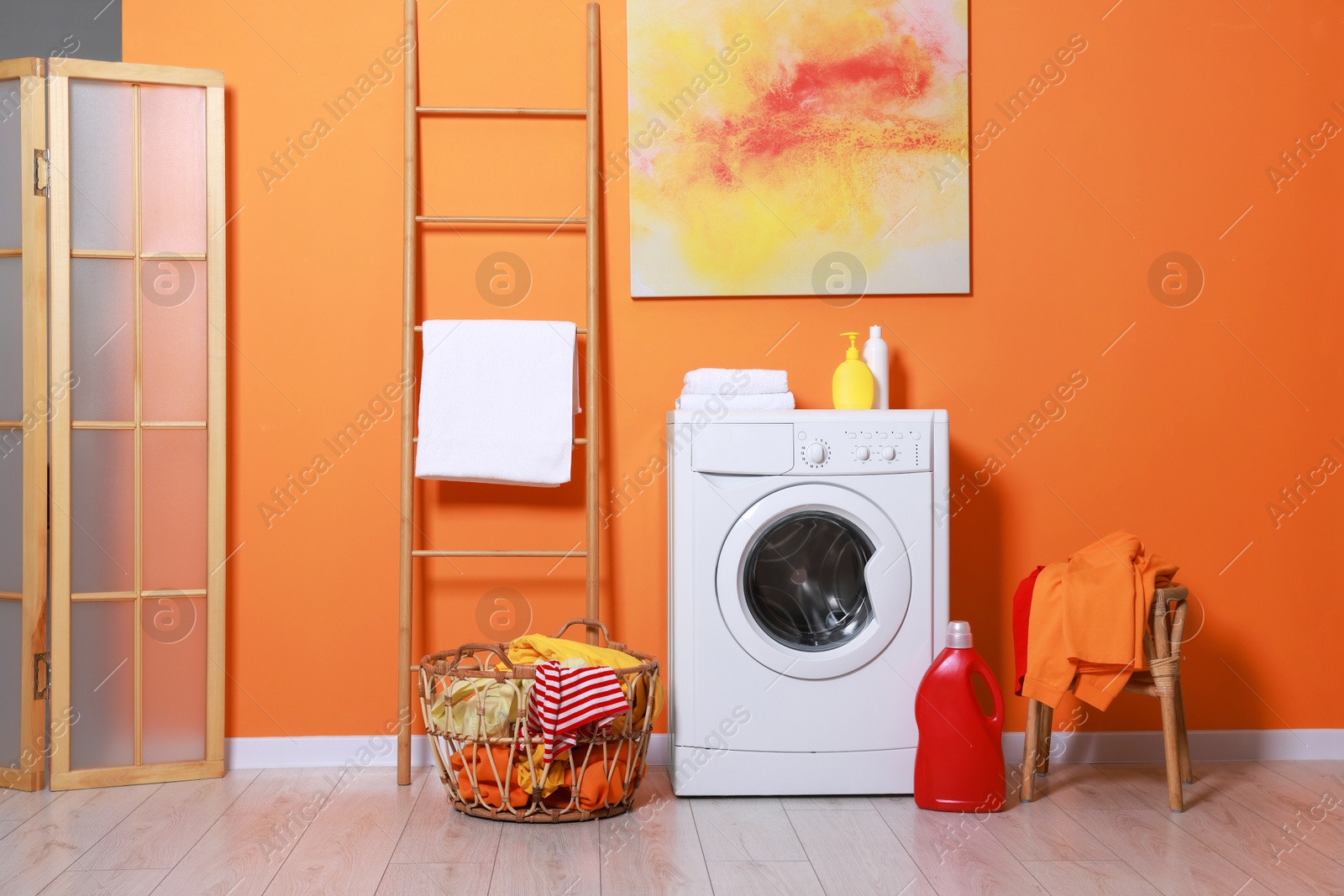 Photo of Washing machine, detergents and basket with laundry near orange wall indoors