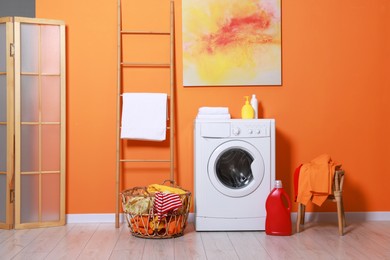 Photo of Washing machine, detergents and basket with laundry near orange wall indoors