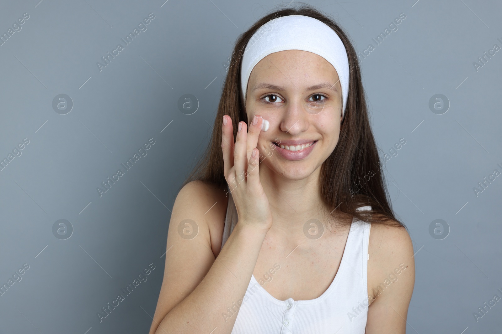 Photo of Teenage girl with acne problem applying cream on grey background