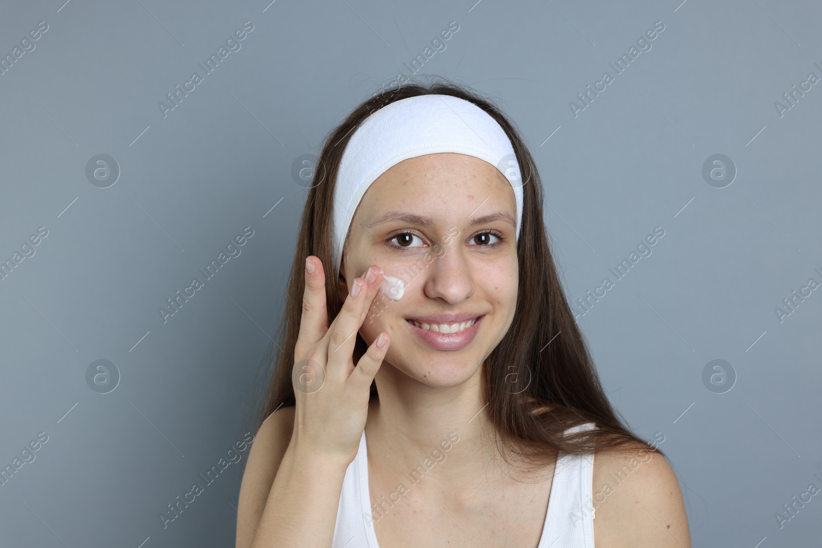 Photo of Teenage girl with acne problem applying cream on grey background