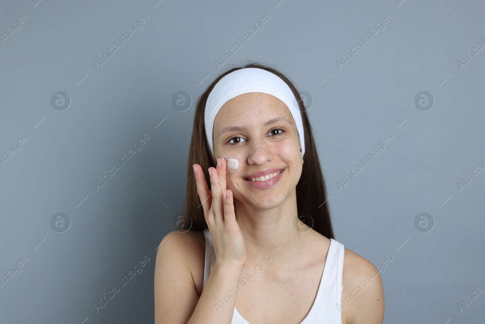 Photo of Teenage girl with acne problem applying cream on grey background