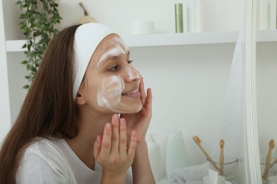 Photo of Teenage girl applying cleansing foam onto her face near mirror indoors. Acne treatment