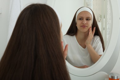 Photo of Upset teenage girl with acne problem looking in mirror indoors