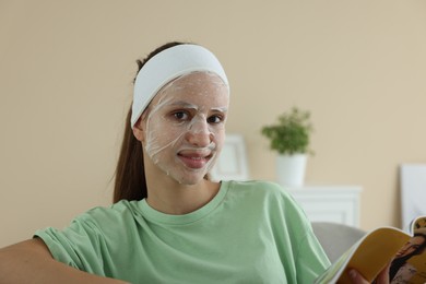 Photo of Teenage girl with sheet facial mask reading magazine at home. Acne treatment