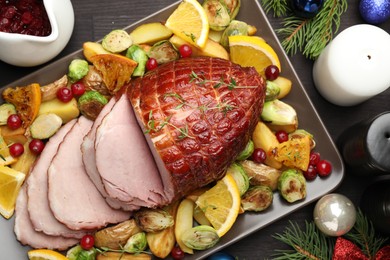 Tasty baked ham served with vegetables and oranges on black wooden table, flat lay