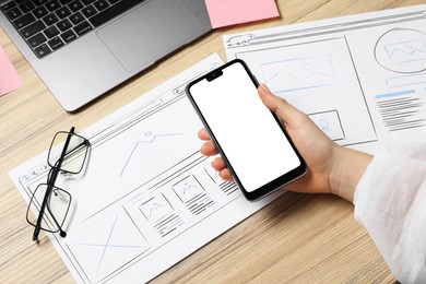 Website designer holding smartphone at wooden table with wireframes and laptop, closeup