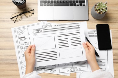 Photo of Website designer holding wireframe at wooden table with smartphone and laptop, top view