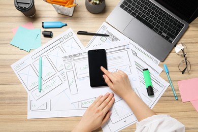 Photo of Website designer using smartphone at wooden table with wireframes, stationery and laptop, top view