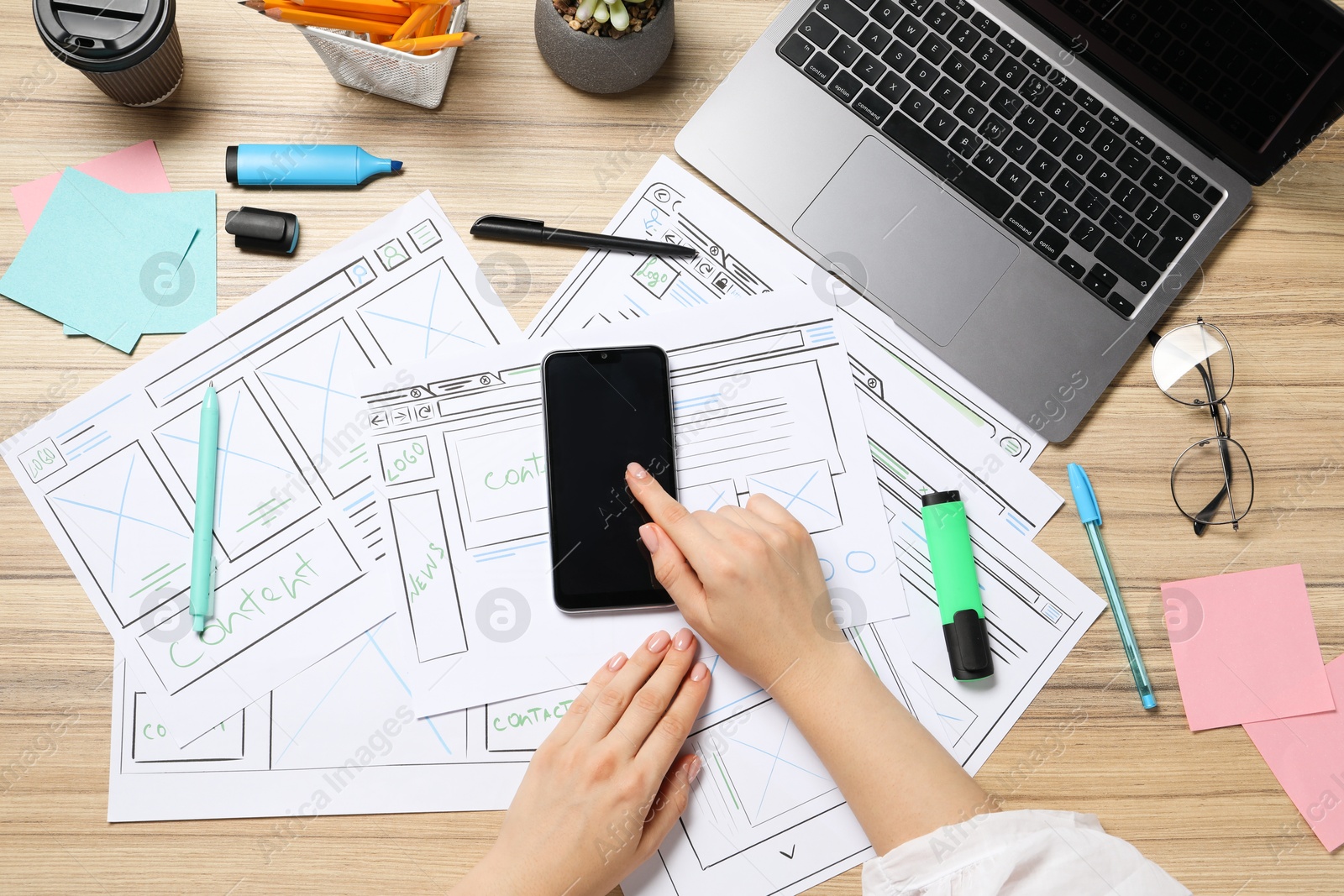 Photo of Website designer using smartphone at wooden table with wireframes, stationery and laptop, top view