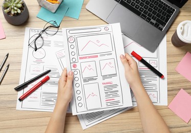 Photo of Website designer holding wireframe at wooden table with stationery and laptop, top view