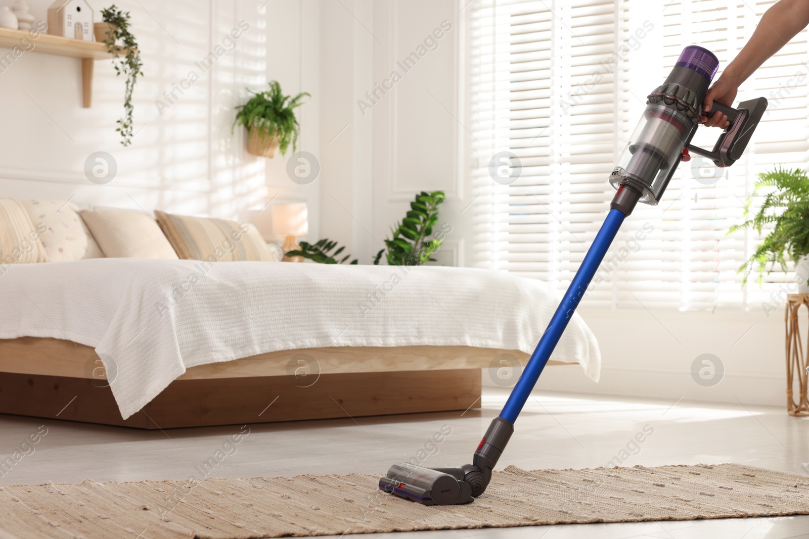 Photo of Woman cleaning rug with cordless vacuum cleaner in bedroom, closeup