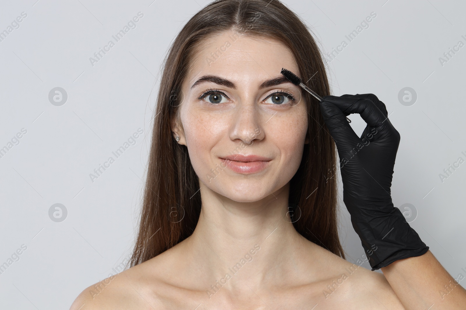 Photo of Beautician brushing client's eyebrows after henna dyeing procedure on light background, closeup