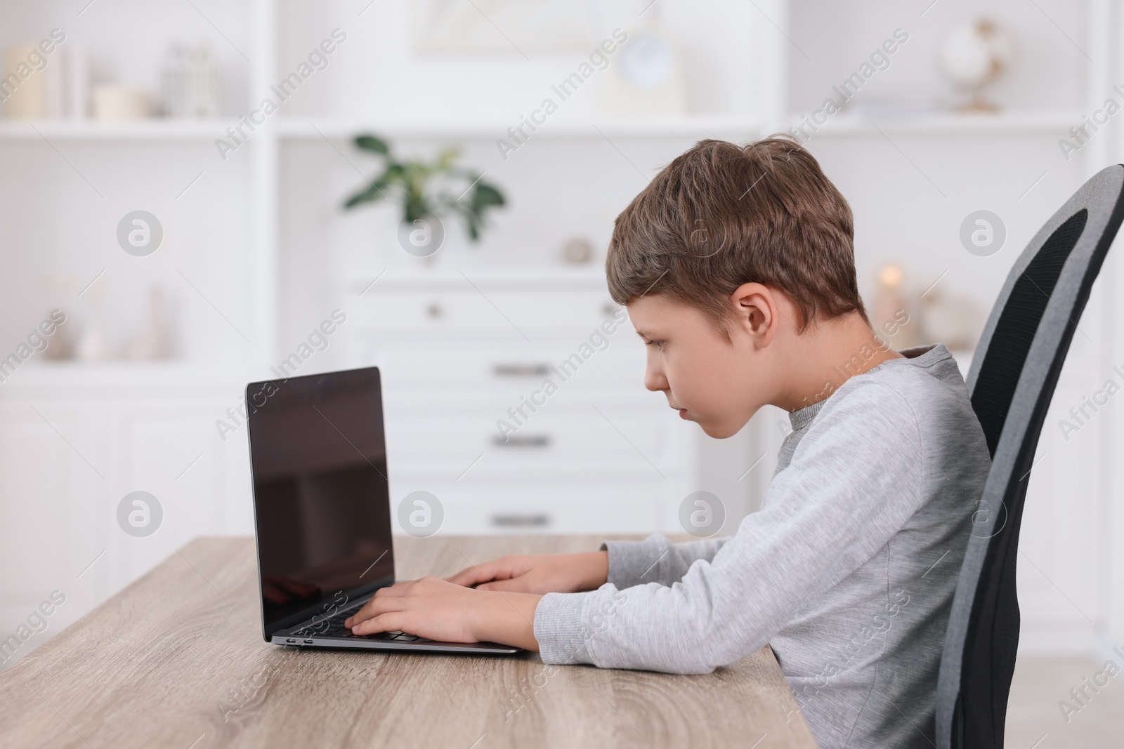 Photo of Boy with incorrect posture using laptop at wooden desk indoors