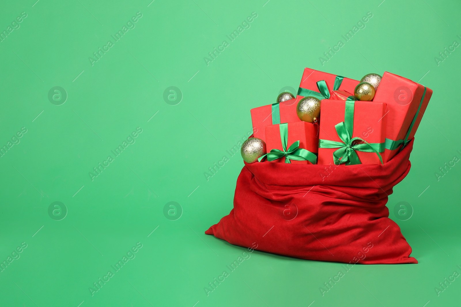 Photo of Red Santa Claus bag with baubles and gift boxes on green background, space for text