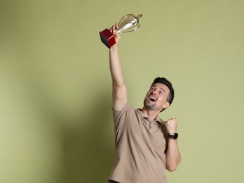 Photo of Happy winner with golden trophy cup on pale olive background, space for text