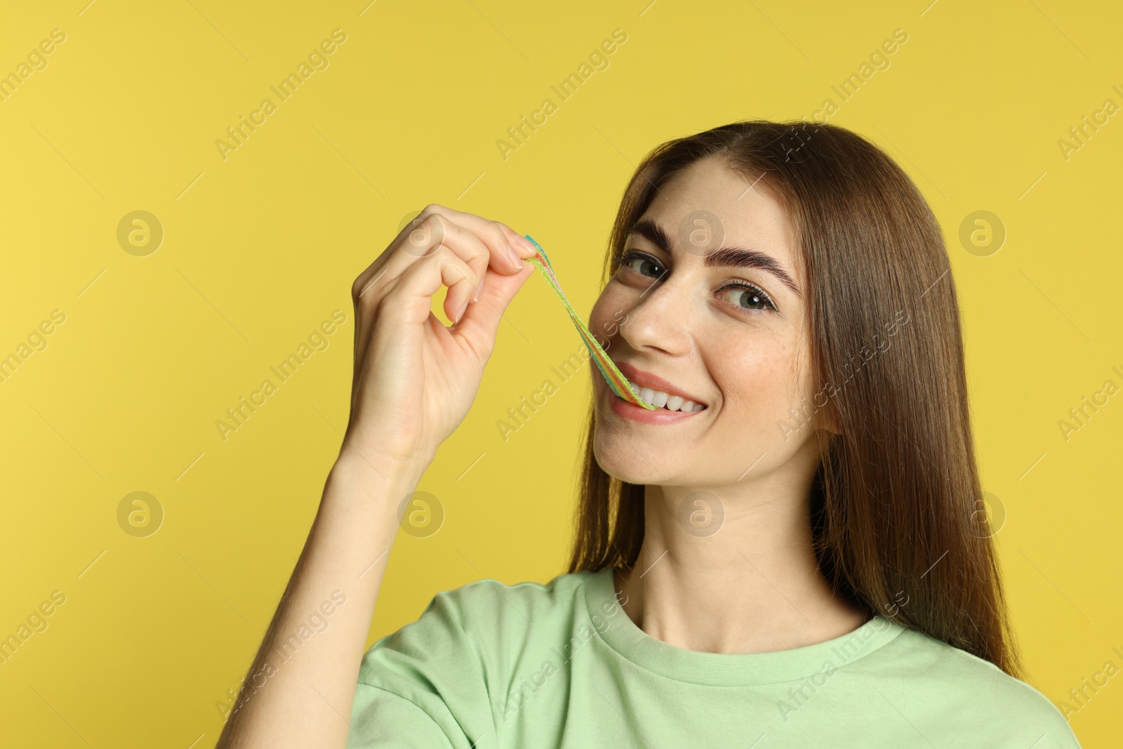 Photo of Young woman eating tasty rainbow sour belt on yellow background