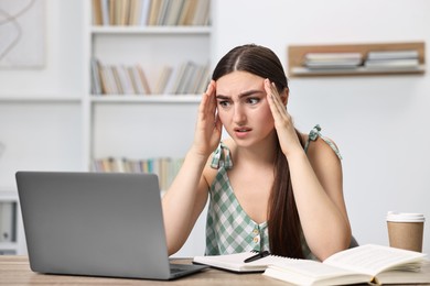 Photo of Tired student preparing for exam with laptop at table indoors