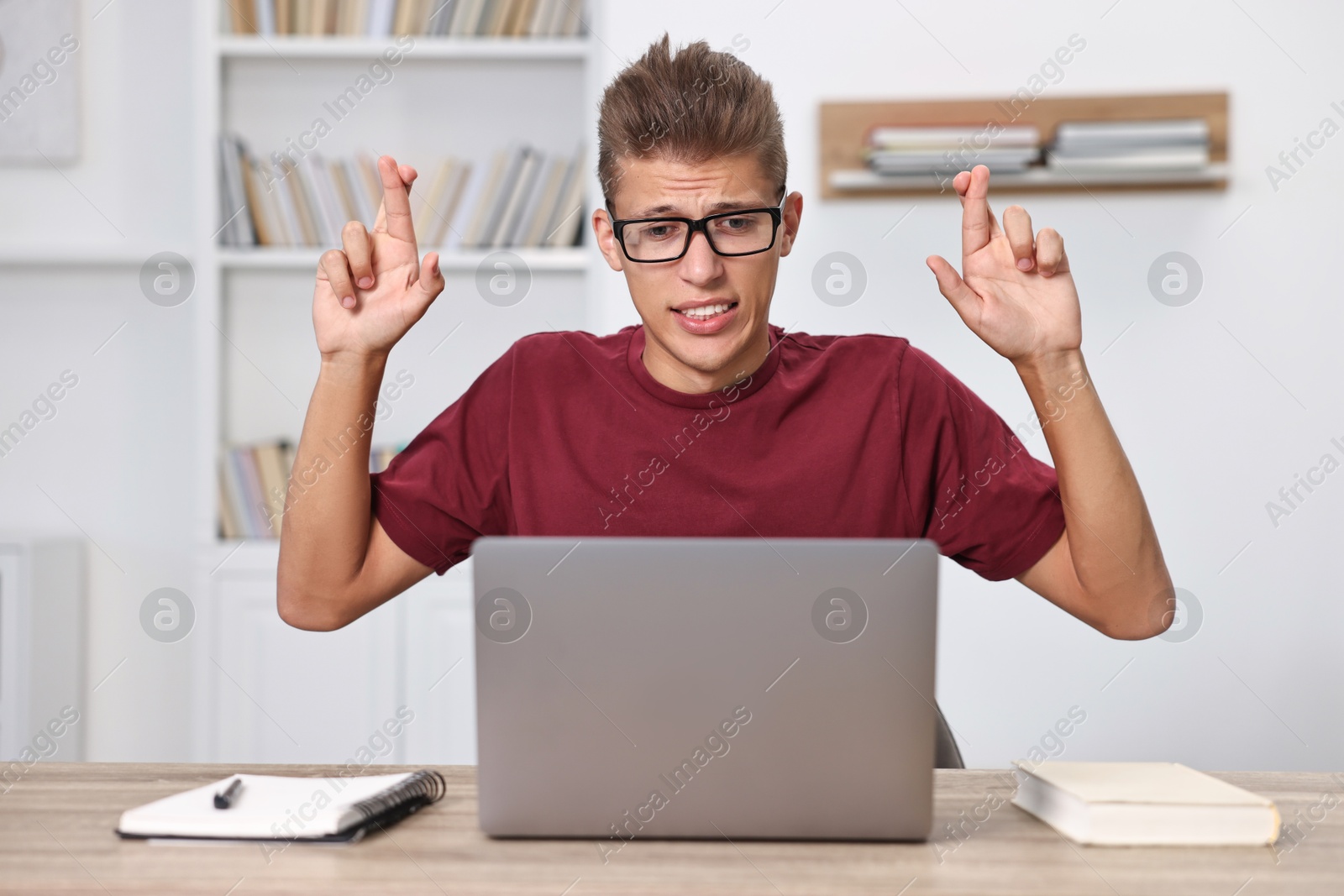 Photo of Worried student with crossed fingers at table with laptop indoors. Hope for good exam result